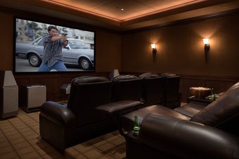 A private home theater with comfortable leather recliners, a large projection screen showing an action movie, and warm ambient lighting from wall sconces and recessed lights.