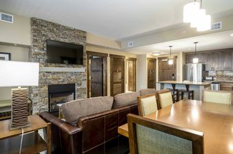 A spacious living room with a stone fireplace, a large leather sofa, a dining table with chairs, and a kitchen area in the background.