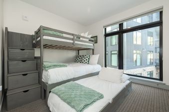 A modern children's bunk room at The Aerie with gray bunk beds, white bedding, green accents, and a large window with an urban view.