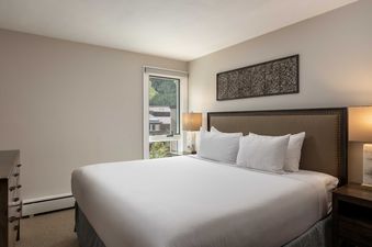 A cozy bedroom at Vail 21 407 features a bed with white linens, an upholstered headboard, and a window showcasing a green hillside view.