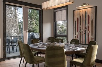 The dining area at Frosthaven Chalet features a large wooden table set for eight, surrounded by olive green velvet chairs, with a view of an outdoor deck and a barbecue grill through sliding glass doors.