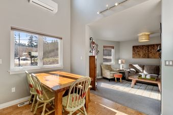 A cozy dining area with a rustic wooden table and light green chairs, illuminated by natural light from large windows overlooking a scenic outdoor view, with a glimpse of the adjacent living room.