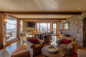 A cozy, luxuriously styled great room in The Developers Residence at See Forever featuring stone walls, wooden beams, comfortable armchairs, a fireplace, and large windows with mountain views.