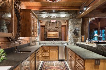 The Royal Wulff Lodge's rustic bar features a U-shaped design with dark countertops and light wood cabinetry, a sink, a compact refrigerator, and a patterned rug, with a stone back wall displaying a cowboy painting, all illuminated by track lighting.