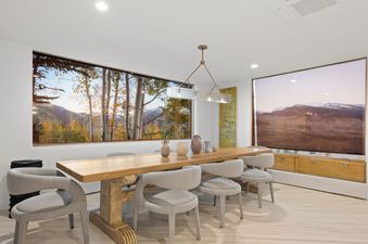The Eppley Estate dining area showcases a large wooden table with six light grey chairs, a modern chandelier, and large windows revealing a vibrant autumn mountain landscape.