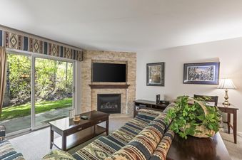 A cozy living room at Snow Flower 135 Park City featuring a comfortable sofa, a dark wooden coffee table, a stone fireplace with a mounted television, and large sliding glass doors opening to a green outdoor view.