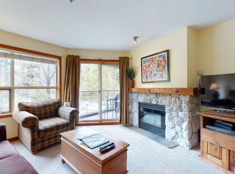 A cozy living room at Aspens on Blackcomb Unit 206 with a stone fireplace, comfortable seating, and a large window offering a view of the natural surroundings.