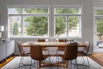 A modern dining area with a wooden table and six brown leather chairs, featuring large windows with views of green trees and mountains, and a light gray rug over hardwood floors.