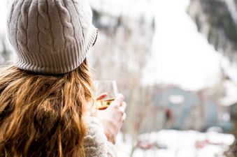 A woman in a white knitted hat and fluffy jacket sips a drink on a balcony overlooking a snowy mountain landscape.