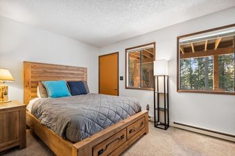 A cozy bedroom in the Grizzly Den with a wooden bed, blue and navy pillows, a rustic nightstand, and windows overlooking a forest.