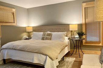 A king-sized bed in a bedroom featuring soft, neutral colors, wooden blinds, and two illuminated bedside lamps.