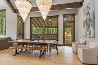 The dining area at Independence Lodge showcases a large wooden table, a bench, and chairs, illuminated by two unique chandeliers, with large windows overlooking an evergreen forest.