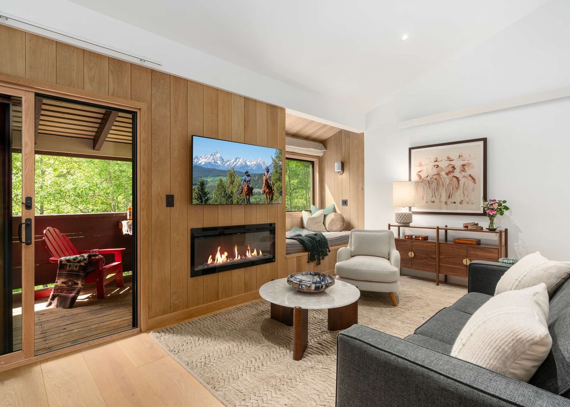 A bright, cozy living room featuring wood paneling, a modern fireplace, a wall-mounted TV, and an open glass door leading to a balcony with forest views.