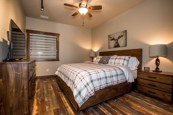 A cozy king bedroom with rustic hardwood floors, a dark wooden bed with plaid bedding, and a moose artwork above the headboard.