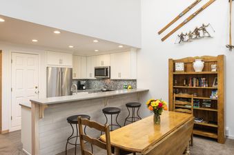 A modern kitchen with white cabinets, stainless steel appliances, a breakfast bar with three stools, and an adjacent dining table with a floral centerpiece.