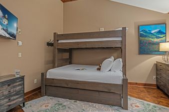A bunk bed with a full-size bed on the bottom and a twin-size bed on top is in a bedroom with a brown dresser with a small lamp on it and a second dresser with a TV on it.