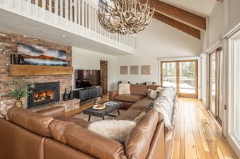 A cozy living room with a large brown leather sectional sofa, a brick fireplace, and a striking antler chandelier, looking out onto a snowy landscape through large windows.