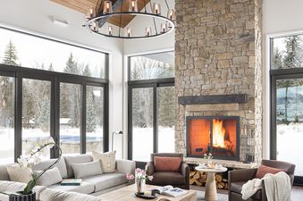 A cozy, spacious living room in a luxury Northern Hideaway home features a lit fireplace, vaulted wooden ceilings, and expansive windows showcasing a snowy outdoor landscape.