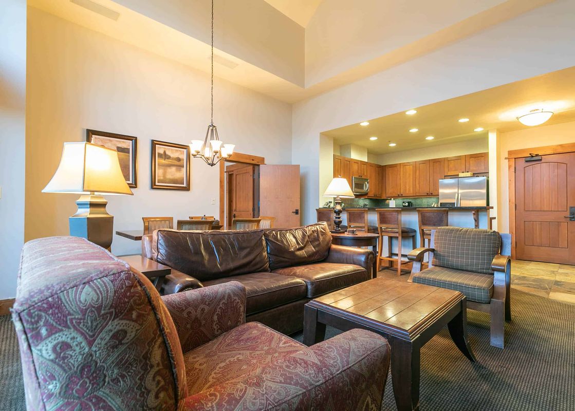A spacious living room with a leather couch, two armchairs, and a wooden coffee table. The room is decorated in a rustic style with wood beams and a chandelier.