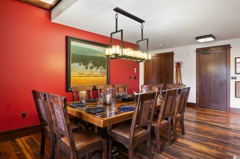 A formal dining area with a long wooden table set with plates and glasses, surrounded by wooden chairs, under a rectangular chandelier, with a red accent wall featuring a large landscape painting.