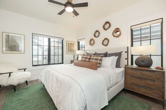 A modern bedroom with a large bed, green rug, and stylish decor, featuring large windows and a ceiling fan.