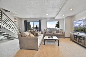The spacious and elegantly furnished lounge at Temptation 7 features two comfortable sofas, a modern coffee table, a large television, and a metal staircase, all set against a backdrop of snowy mountain views.