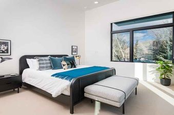 A bedroom with a bed, a nightstand, a lamp, a window with a view of trees, and a bench at the foot of the bed.