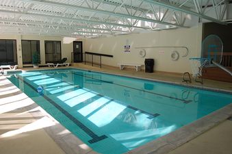 An indoor swimming pool with blue water and white lane lines is well-lit by natural light streaming through large windows, with lounging chairs and safety equipment visible around the edges.