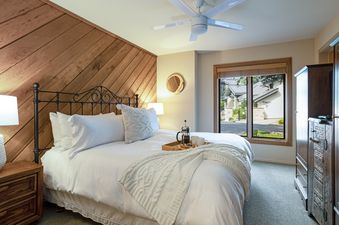 The bedroom at 88 Ridge Point features a large bed with a black wrought iron headboard and white bedding, with a wooden accent wall, a ceiling fan, and a view of the neighborhood through a large window.