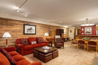 A cozy living room at Montaneros 212 with a vibrant red-orange sectional sofa, a wooden coffee table, and an adjacent dining area with a rustic table and chairs.