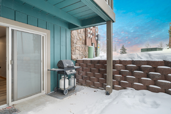 A black barbecue grill sits on a snowy patio outside a building with teal siding and a brown stone wall.