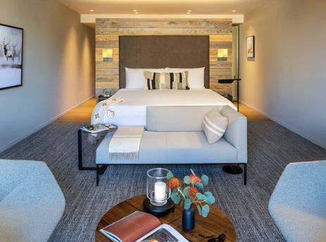 A modern bedroom with a king-sized bed, a chaise lounge, two armchairs, and a large artwork of deer in a snowy landscape.