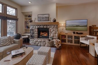 A cozy living room with a lit fireplace, a large TV, comfortable sofas, and a large window showing a snowy outdoor view.