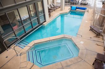 An aerial view of the outdoor pool and hot tub at Chateau Chamonix CX326, featuring clear blue water, a surrounding concrete deck with lounge chairs, and an adjacent building with large windows.