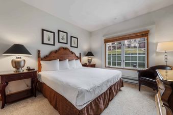 A cozy bedroom with a large bed, wooden headboard, bedside lamps, botanical prints, and a window with an outdoor view.