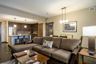 The Trailhead Lodge 4115 living room features a sectional sofa with a large pillow, a coffee table with books and remote controls, and a large window that overlooks the surrounding area.