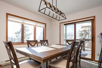 A bright dining room with a large wooden table, chairs, and an industrial-style chandelier, offering views of a snowy landscape through expansive windows.