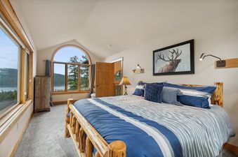 A cozy bedroom with a rustic log bed, blue and white striped bedding, and large windows offering a scenic view of a lake and forest, alongside an elk print on the wall.