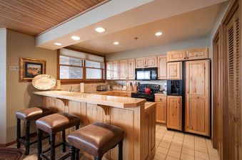 A fully equipped kitchen with natural wood cabinets, black appliances, a breakfast bar with leather stools, and tiled floors at Chateau Chamonix CX124.