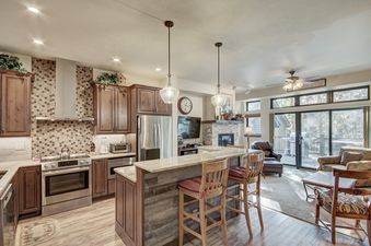 A modern kitchen with a stainless steel stove and a breakfast bar that overlooks a living room with a fireplace and large windows.