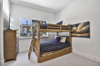 A modern bedroom features light wood bunk beds with dark blue plaid bedding, a matching dresser with a television, and a large window overlooking an outdoor area with other buildings.