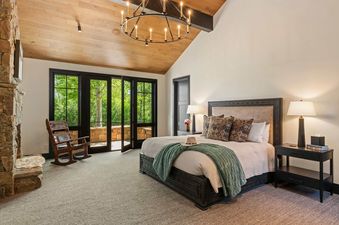 A luxurious master bedroom at Teton Crest with a vaulted ceiling, stone fireplace, and double glass doors leading to a leafy outdoor balcony.