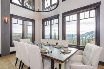 An elegant dining room at 7 Lower Cascade Ridge features a dark wood table set for eight, upholstered chairs, and a large chandelier, all against a backdrop of snowy mountain views.