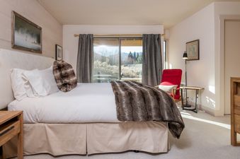 A bedroom with a large bed, white linens, a furry throw, a bedside table, and a view of a mountain range outside the window.