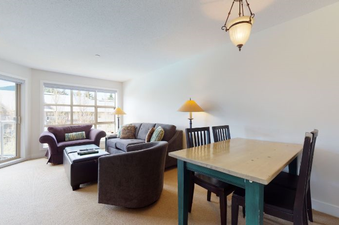 A cozy living space with a dining area features a plush purple sofa, a comfortable gray sofa, and a brown armchair, all arranged around a dark ottoman on light-colored carpet, with natural light streaming in from large windows offering a view of the outdo