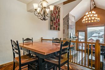 A large wooden dining table with black chairs is set in the Hygge Hideaway, with a cozy fireplace and expansive window view of snow-covered mountains in the background.