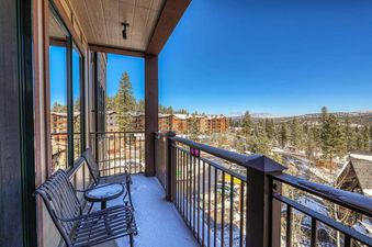 A snow-covered balcony with two metal chairs and a small table overlooking a snowy mountainside with evergreen trees.