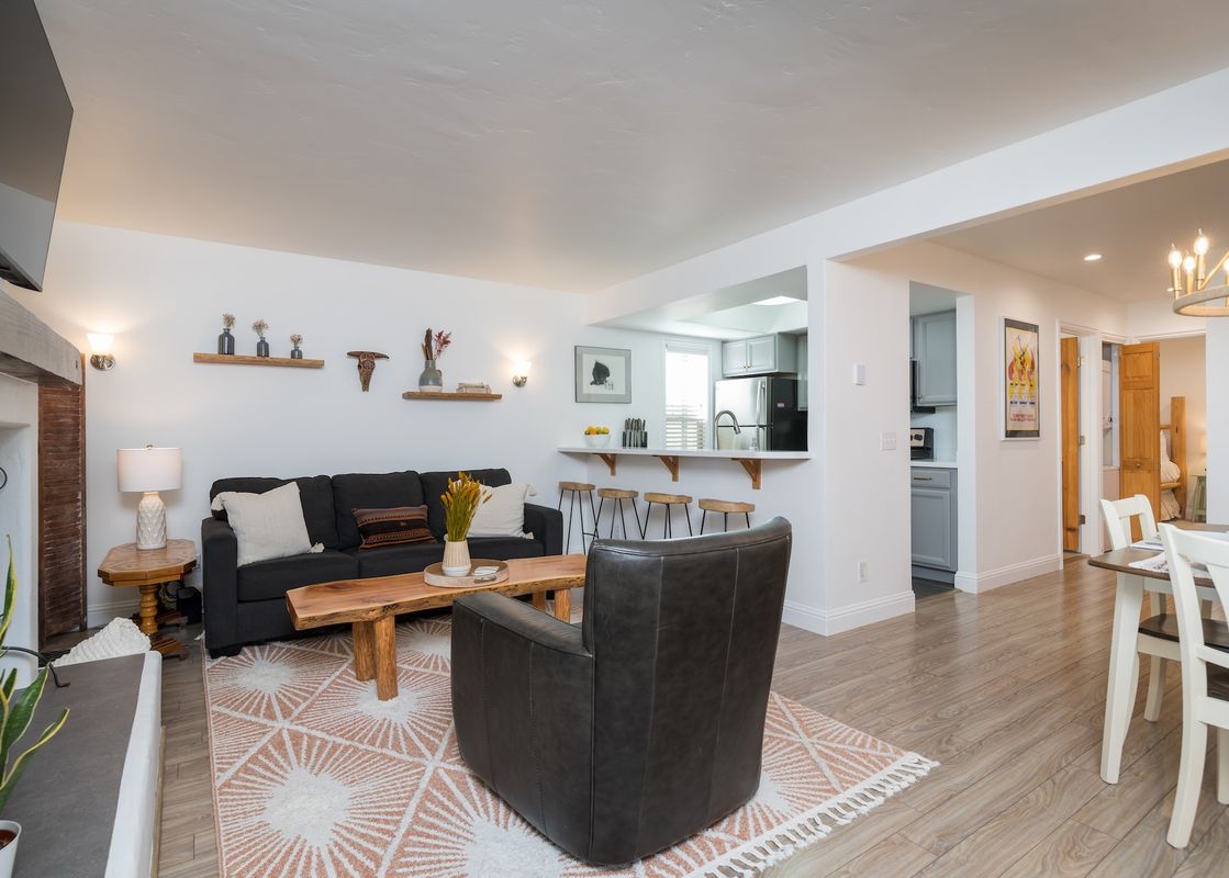 A stylish open-concept living area with a dark gray sofa, leather armchair, live-edge coffee table, and a breakfast bar leading to a modern kitchen.