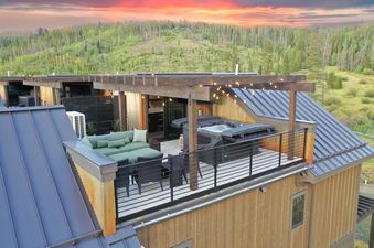 An elevated view of Sky High Colorado's rooftop deck at sunset, featuring an inviting hot tub, comfortable outdoor seating, and string lights, all set against a backdrop of forested mountains and a vibrant sky.