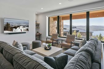 A luxurious living room at 11981 N Gemini Way SkyRidge, featuring a gray sectional sofa, elegant armchairs, and a large flat-screen TV, all set against a backdrop of expansive windows showcasing a scenic lake and mountain view.